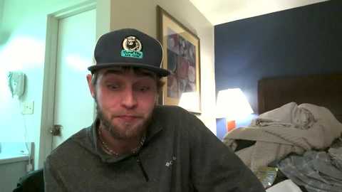 Media: Video of a bearded man in a dark baseball cap, gray hoodie, and necklace, sitting on a bed with messy sheets in a dimly lit room.