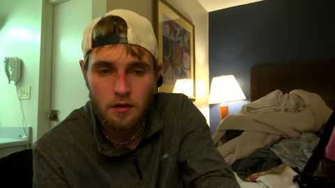 Media: Video of a bearded man in a grey shirt and backwards baseball cap, sitting in a dimly-lit hotel room with disheveled bed, bedside lamps, and framed art on the wall.