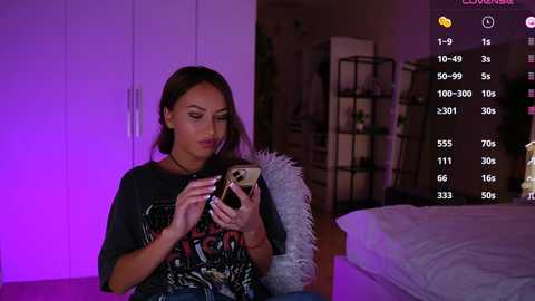 Media: A video of a woman with straight brown hair, wearing a dark graphic t-shirt, sitting on a white chair in a dimly lit room with purple lighting, holding a phone.