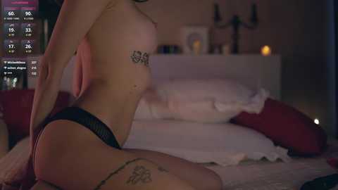 A video of a topless woman in black panties, kneeling on a bed with red pillows. She has a tattoo on her left breast and another on her thigh. The background shows a dimly lit room with a TV screen displaying a video game.