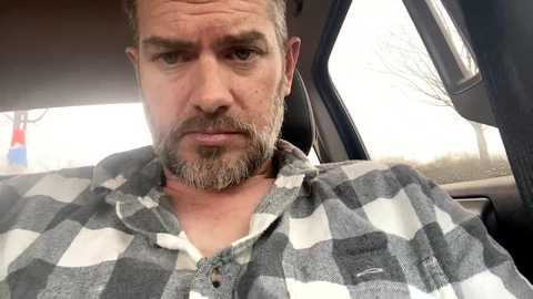 Media: Video of a middle-aged man with a short beard and mustache, wearing a grey and white plaid shirt, sitting in a car. Background shows blurry trees and a traffic cone.