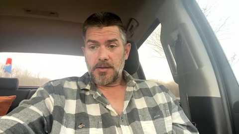 Media: Video of a bearded man with short brown hair wearing a gray and white plaid shirt, sitting in the driver's seat of a car with a neutral expression.