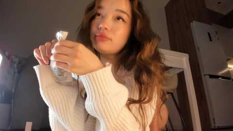 Media: Video of a young Asian woman with long, wavy brown hair, wearing a white, ribbed sweater, holding a silver bottle. The background shows a dimly lit kitchen with a white refrigerator and wooden cabinets.