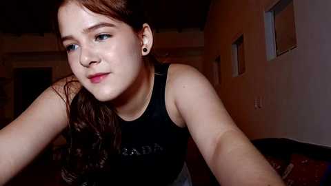 Media: Video of a young woman with fair skin, brown hair, and blue eyes, wearing a black sleeveless top, in a dimly lit room with beige walls and windows.