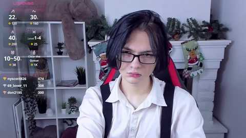 Media: Video of a young Asian man with long black hair, wearing glasses, a white shirt, and black suspenders, standing in a cozy living room with a Christmas tree, plants, and shelves.