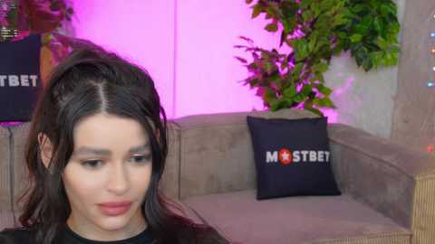 Media: Video of a young woman with long, dark hair, wearing a black top, sitting on a beige couch with a \"Mister Bet\" pillow. The background features green plants and a pink-lit wall.