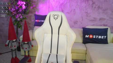 Media: Video of a gaming setup with a white gaming chair, decorated with \"MSI\" logo, positioned in front of a beige sectional sofa. Two plush toy robots and floral decor adorn the background.