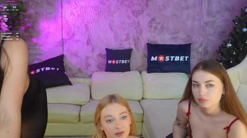 Media: Video of two young women with pale skin and long hair, wearing red lingerie, sitting on a cream couch. Background features purple lighting, textured wall, and dark pillows with \"MSTBET\" text.