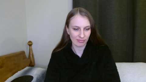 Media: Video of a Caucasian woman with long, straight brown hair, fair skin, and a slim build, wearing a black coat, sitting on a bed with a wooden headboard and grey curtains in the background.