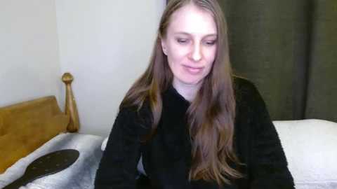 Media: Video of a fair-skinned, long-haired Caucasian woman in a black sweater, sitting on a bed with white sheets and a wooden headboard, against a beige wall with a dark curtain.