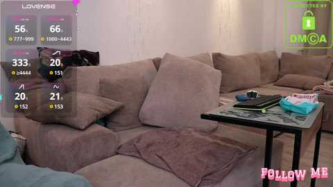 Media: Video of a cozy living room with a large beige sectional sofa, scattered cushions, a black coffee table, and a gaming monitor displaying a 'LOVECRAFT' game screen.