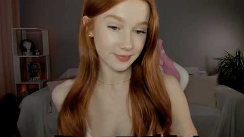 Video of a young woman with long, straight red hair, fair skin, and small breasts, wearing a light-colored top, in a dimly lit bedroom with gray walls, a white shelf, and a pink cushion.