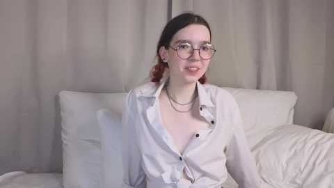 Media: Video of a young, fair-skinned woman with glasses and red hair, wearing a loose white shirt, seated on a beige couch with white pillows.