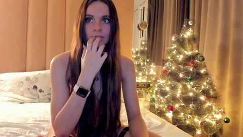 Media: Video of a slender, pale-skinned woman with long brown hair, wearing a black tank top and smartwatch, sitting on a bed with a festive, lit-up Christmas tree in the background.