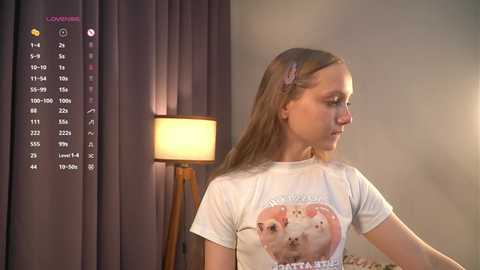 Media: Video of a young woman with long blonde hair, wearing a white t-shirt with a pink rabbit graphic, standing beside a lit lamp in a cozy room with grey curtains.