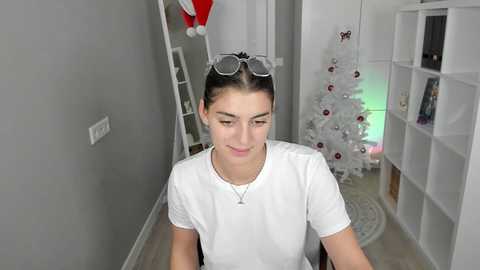 Media: A video of a young woman with dark hair in a bun, wearing a white T-shirt, clear glasses, and a necklace, sitting in a modern living room with a white Christmas tree, white shelves, and a red Santa hat.