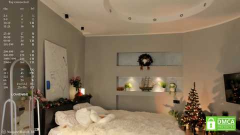 Media: A video of a cozy, modern bedroom with a cream-colored bed, white pillows, a small Christmas tree, and a wreath on the wall. The room features recessed shelves, soft lighting, and a minimalist decor style.