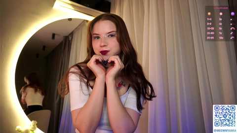 Media: Video of a fair-skinned, long-haired brunette woman with red lipstick, wearing a white top, holding her hands in a heart shape in front of a makeup mirror.