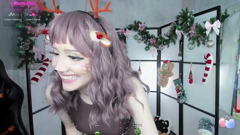 Media: Video of a smiling woman with wavy purple hair, wearing reindeer antlers and a black top, surrounded by festive Christmas decorations in a well-lit room.