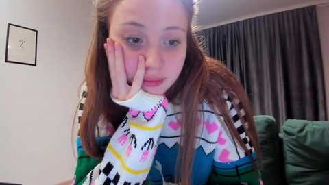 Media: Video of a young Caucasian girl with long brown hair, wearing a colorful, patterned sweater, resting her head on her hand, in a cozy living room with green couches and gray curtains.