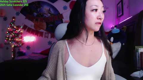 Media: Video of a young woman with long black hair, wearing a white tank top and beige cardigan, in a festive, colorful bedroom with holiday decorations.