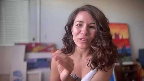 Media: Video of a middle-aged woman with long, wavy brown hair, wearing a white tank top, speaking animatedly in a home studio with colorful paintings and shelves in the background.