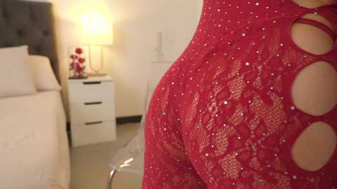 Media: Video of a woman in a red lace dress with cut-outs, standing in a modern bedroom with white furniture, a lamp, and a bed.