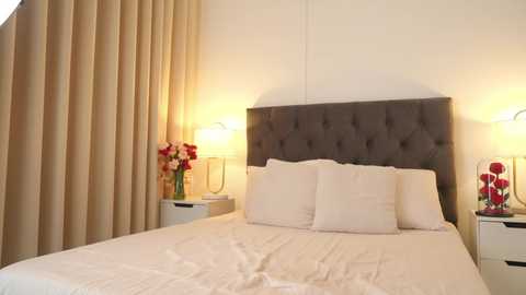 Media: Video of a modern bedroom with beige curtains, a gray upholstered headboard, white linens, two pillows, a vase of red flowers on a white nightstand, and a lamp.