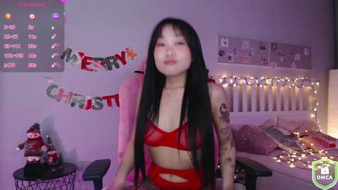 Media: A video of an Asian woman with long black hair and tattoos, wearing a sheer red bra and panties, in a Christmas-themed bedroom with a bed, ornaments, and a \"Merry Christmas\" banner.