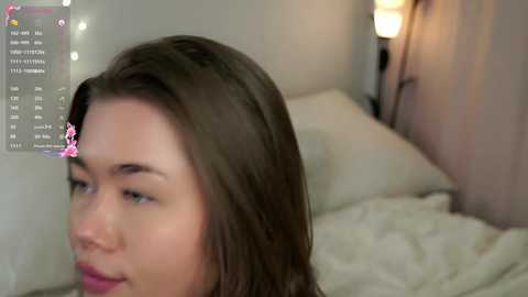 Media: Video of a Caucasian woman with shoulder-length brown hair, adorned with a pink flower clip, gazing thoughtfully at a screen displaying text. Background features a bed with white linens, a nightstand lamp, and a wall-mounted string of fairy lights.