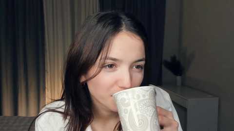 Media: Video of a young woman with long dark hair, fair skin, and brown eyes, wearing a white top, sipping from a white ceramic mug with intricate designs, in a dimly lit bedroom with dark curtains and a white dresser in the background.