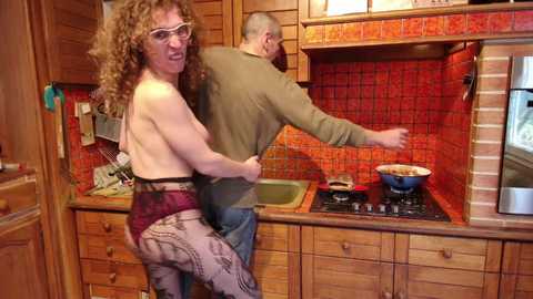 Media: Video of a curly-haired, topless woman in a maroon skirt, embracing a bald man in a green shirt, cooking in a rustic kitchen with red tiled backsplash and wooden cabinets.