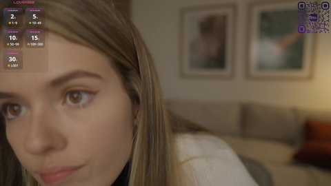 Media: Video of a young woman with long, straight blonde hair, light skin, and brown eyes. She wears a black top and looks thoughtful. Background features a beige sofa, framed pictures, and a soft, warm ambiance.