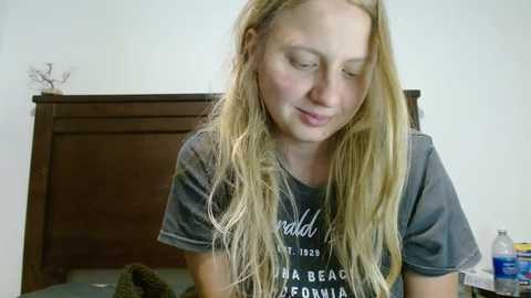 Media: A video of a young, fair-skinned blonde woman with long, wavy hair, wearing a grey T-shirt, sitting on a wooden bed with a dark headboard. She appears to be daydreaming.