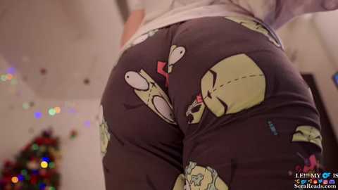 Video of a person's lower back and buttocks wearing cartoon-themed pajama pants featuring a pink heart and three cartoonish faces with exaggerated expressions. The background shows festive, blurred lights and a decorated Christmas tree.