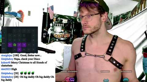 Media: A video of a shirtless, bearded, red-haired man with glasses and a leather harness, standing in a brightly lit, cluttered room with a camera in front. Text overlays the image with chat messages and a live stream title.