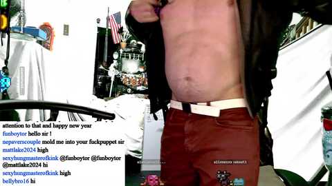 Media: Video of a shirtless man in brown pants and a black jacket, standing in a cluttered room with American flags, a car, and text overlay.