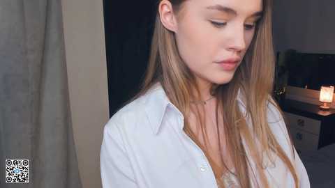 Media: Video of a young woman with fair skin, long blonde hair, wearing a white button-down shirt, standing indoors near a lit candle on a nightstand.