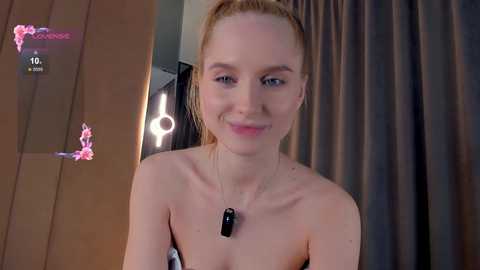 Media: Video of a fair-skinned, blonde woman with a thin frame, wearing a strapless top and a black pendant necklace. She smiles in a modern room with a mirror and dark curtains.