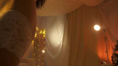 Media: Video of a dimly lit, cozy room with warm lighting, featuring a white lace curtain, a golden candle, and a soft, yellow glow from a nearby lamp.