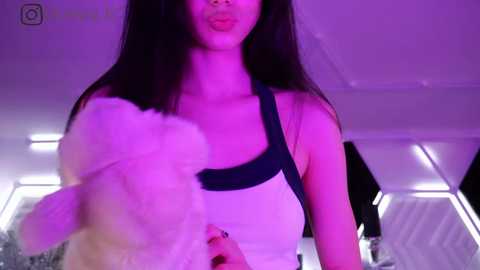 Media: Video of a young woman with long black hair, wearing a white tank top with a black strap, blowing a bubble with a pink bubble gum. Background is a modern room with purple lighting and white walls.