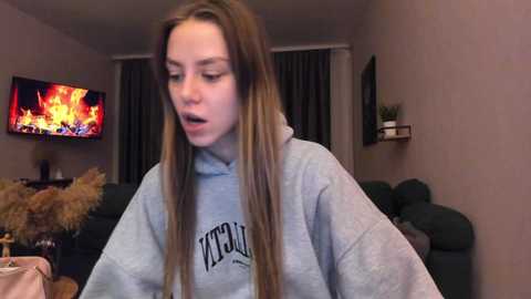 Media: Video of a young woman with long, straight brown hair, wearing a light gray hoodie, standing in a cozy living room with a TV displaying a fire, a green couch, and a fireplace in the background.