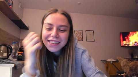 Media: Video of a young woman with straight brown hair, wearing a gray sweater, laughing with braces, in a cozy living room with a fireplace, shelves, and framed art on the wall.