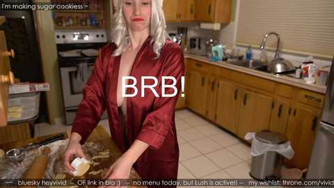 Media: A video shows a woman with platinum blonde hair, wearing a red satin robe, baking in a modern kitchen with wooden cabinets. Text overlays include \"I'm making sugar cookies\" and \"BB!\".