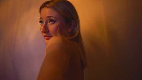 Media: Video of a nude, fair-skinned woman with shoulder-length blonde hair, gazing contemplatively to the side. Warm, soft lighting creates a gentle glow on her smooth skin. Background is a blurred gradient of warm colors, enhancing the intimate, serene atmosphere.