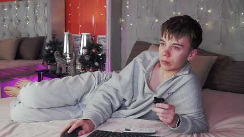 Media: Video of a young man with short brown hair, wearing gray pajamas, lying on a bed, holding a remote, in a cozy, warmly lit bedroom with Christmas decorations.