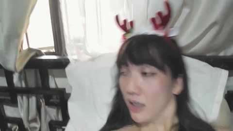 Media: A video of a young East Asian woman with long black hair, wearing a red and white reindeer headband, lying on a white bed with light streaming through the window.