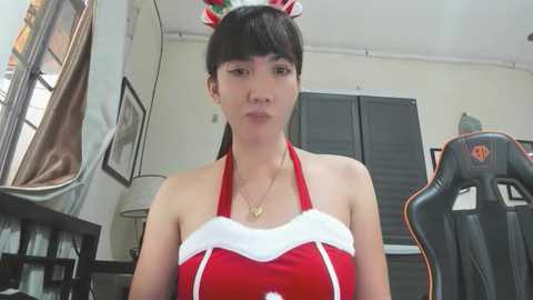 Media: Video of a young Asian woman in a red Santa-themed dress with a white collar, wearing a red Santa hat. She sits in a gaming chair in a cluttered room with framed pictures and a window.