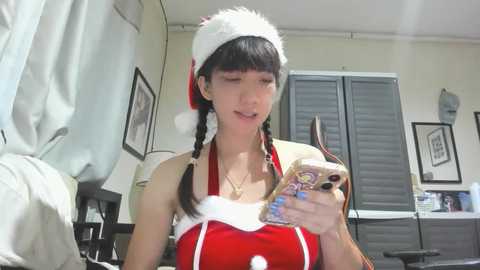 Media: A video of an Asian woman in a red Santa dress with a white fur trim, holding a phone, standing in a home with framed pictures and a window with closed blinds in the background.