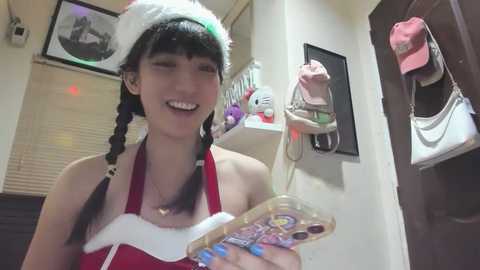 Media: Video of a smiling Asian woman with long braided black hair, wearing a red Santa hat and apron, holding a festive candy box. Background features framed photos and stuffed toys.